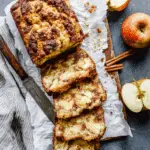 Cinnamon Apple Bread loaf with slices showing apple chunks and cinnamon swirl, served on parchment with fresh apples.