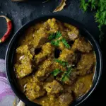 Top view of Coconut Beef Curry in a black bowl, garnished with coriander leaves and surrounded by red onions, garlic, and rice.