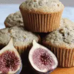 Freshly baked fig muffins stacked on a wooden surface with a ripe fig sliced in half in the foreground.