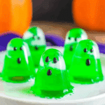 A plate of bright green ghost jello shots shaped like spooky little ghosts with black candy eyes and mouths.