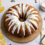 Lemon Zucchini Cake with white glaze and lemon zest, displayed on a wooden board with lemons and herbs around.