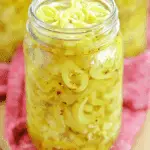 Jar of pickled banana peppers in brine with red pepper flakes, set on a red cloth.