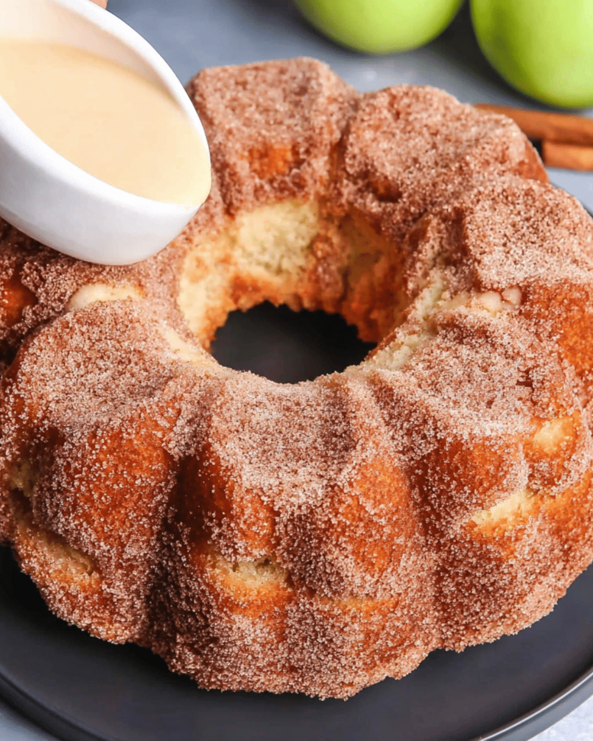 Apple cider donut cake covered in cinnamon sugar, presented on a black plate with a creamy glaze being poured over the top.