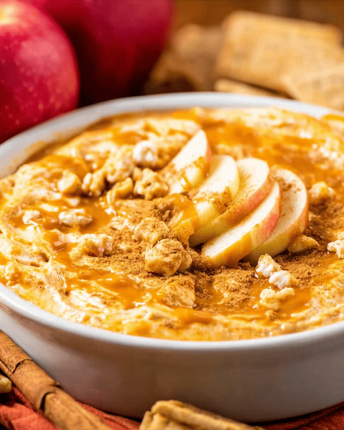 Caramel apple cheesecake dip in a white bowl, topped with fresh apple slices, caramel drizzle, cinnamon, and crumbled graham crackers.