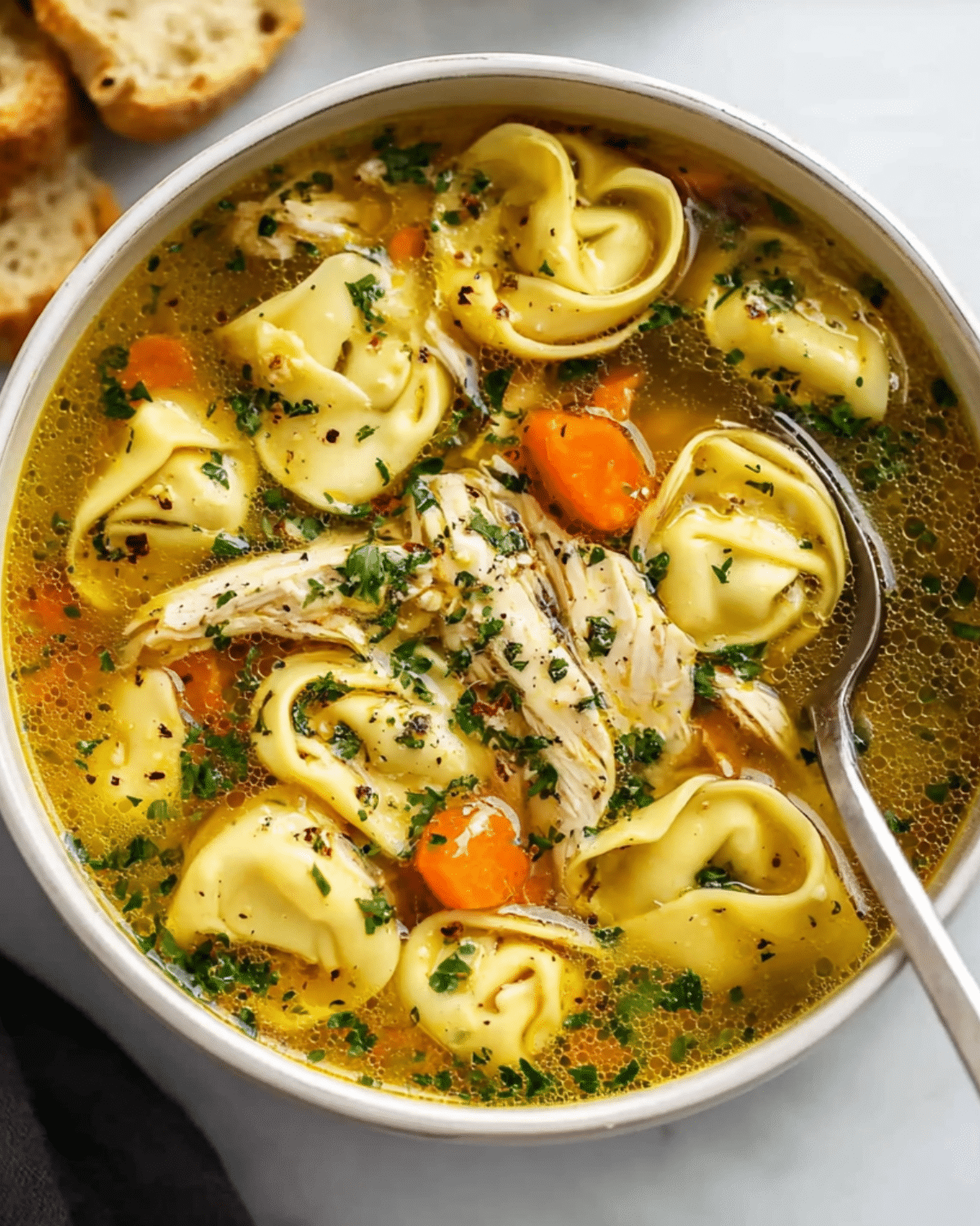 Chicken tortellini soup in a white bowl, filled with cheese tortellini, shredded chicken, carrots, herbs, and a golden broth.