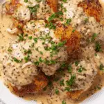 Golden seared chicken thighs covered in a creamy French wine and mustard sauce, garnished with fresh parsley on a white plate.
