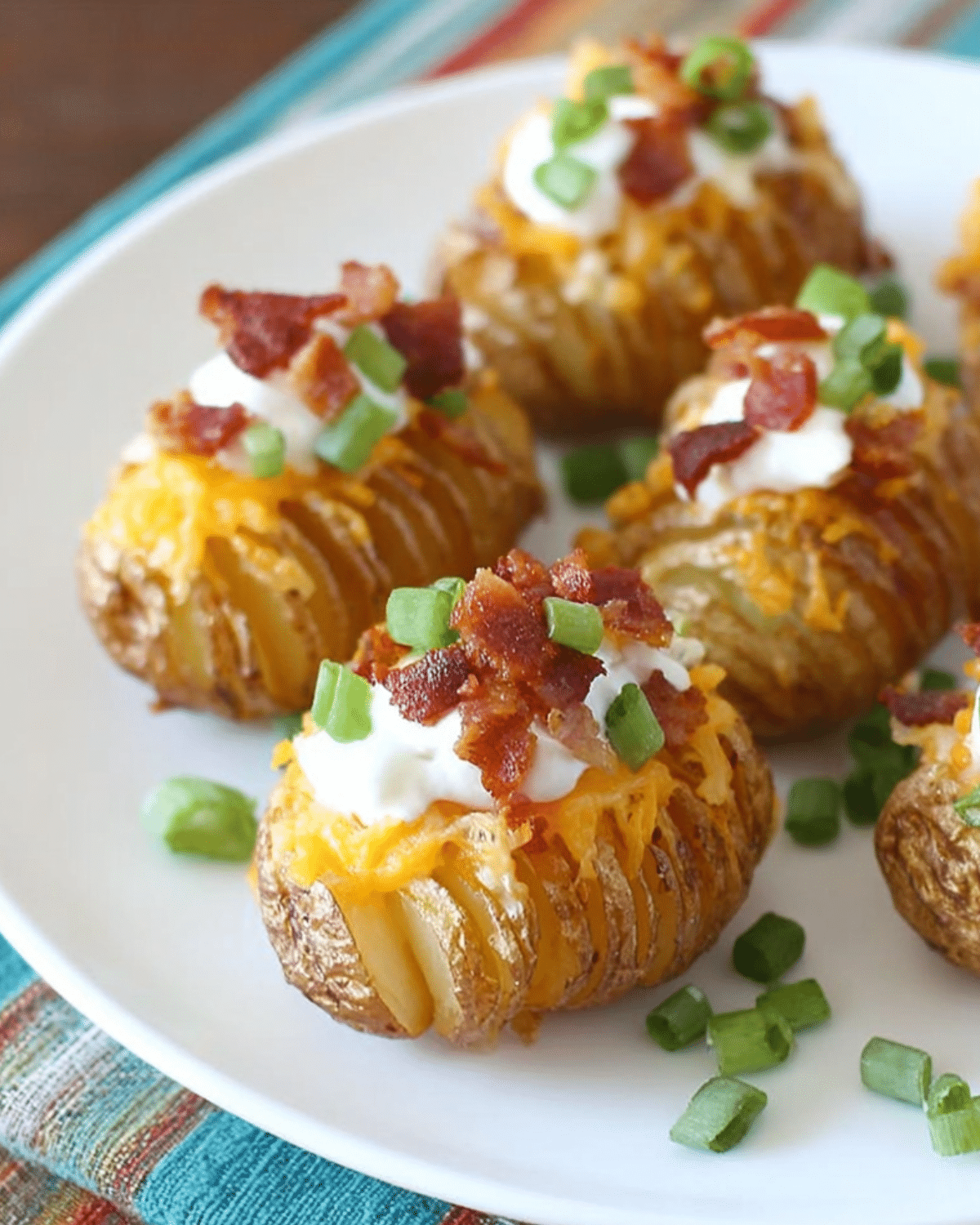 Crispy loaded Hasselback potato bites topped with cheese, sour cream, bacon, and green onions on a white plate.