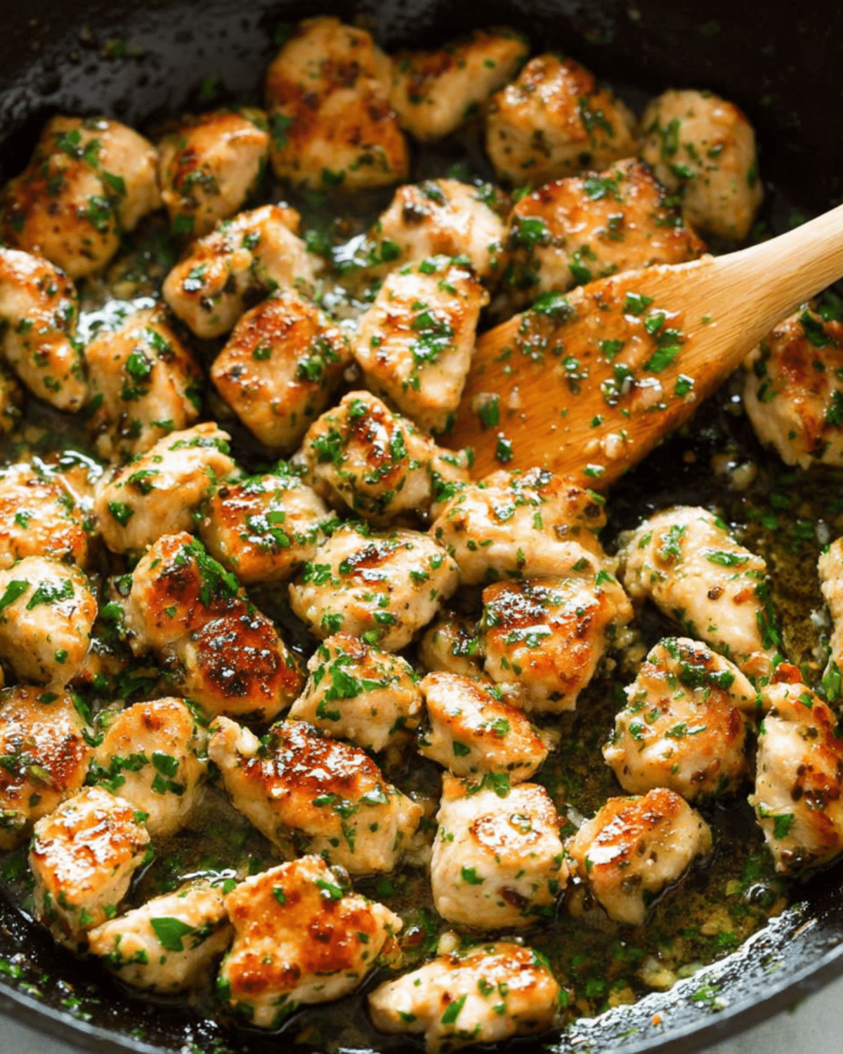 Skillet filled with golden-brown garlic butter chicken bites garnished with chopped parsley and stirred with a wooden spoon.