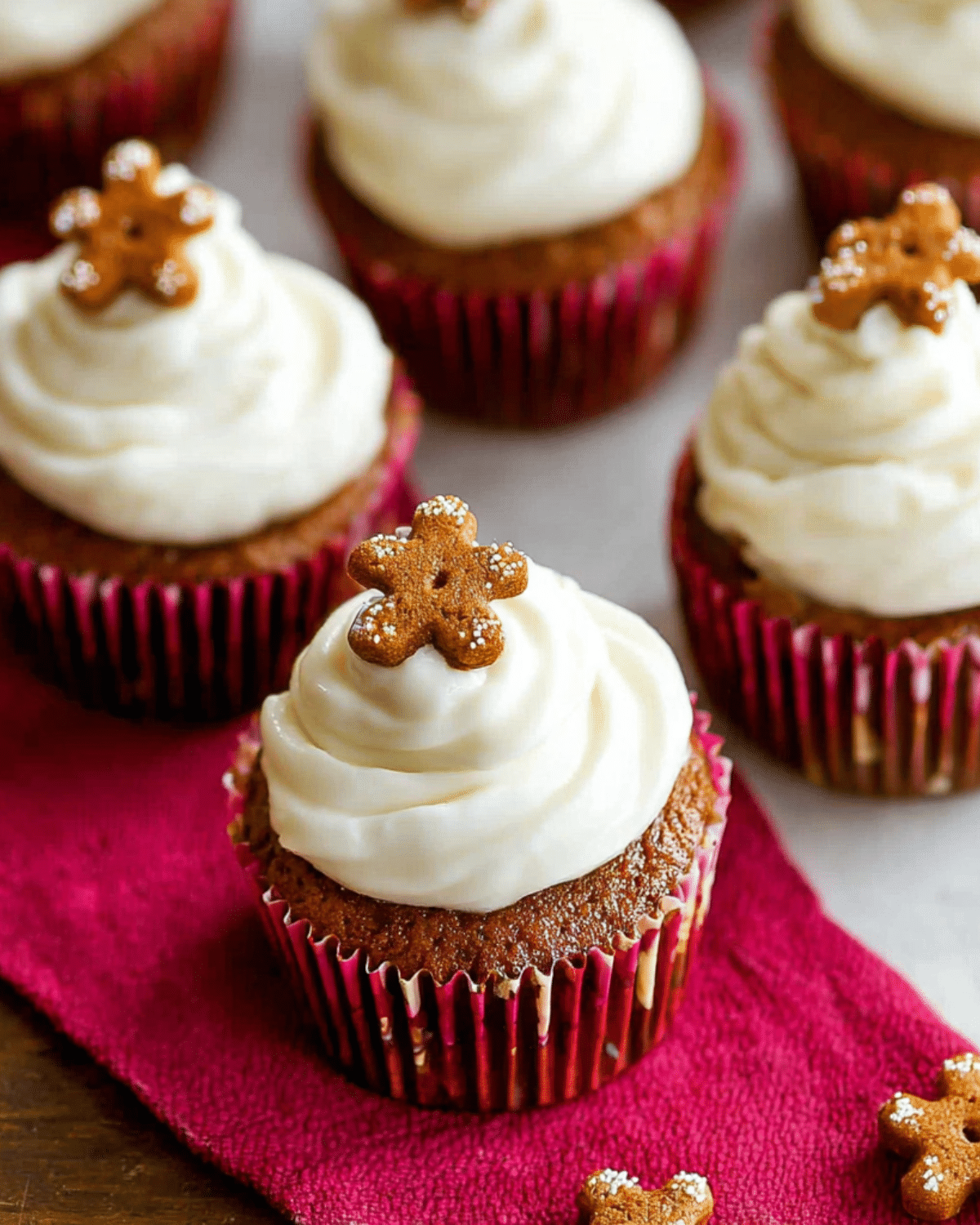 Gingerbread cupcakes with white frosting, each topped with a mini gingerbread cookie, in red cupcake liners.