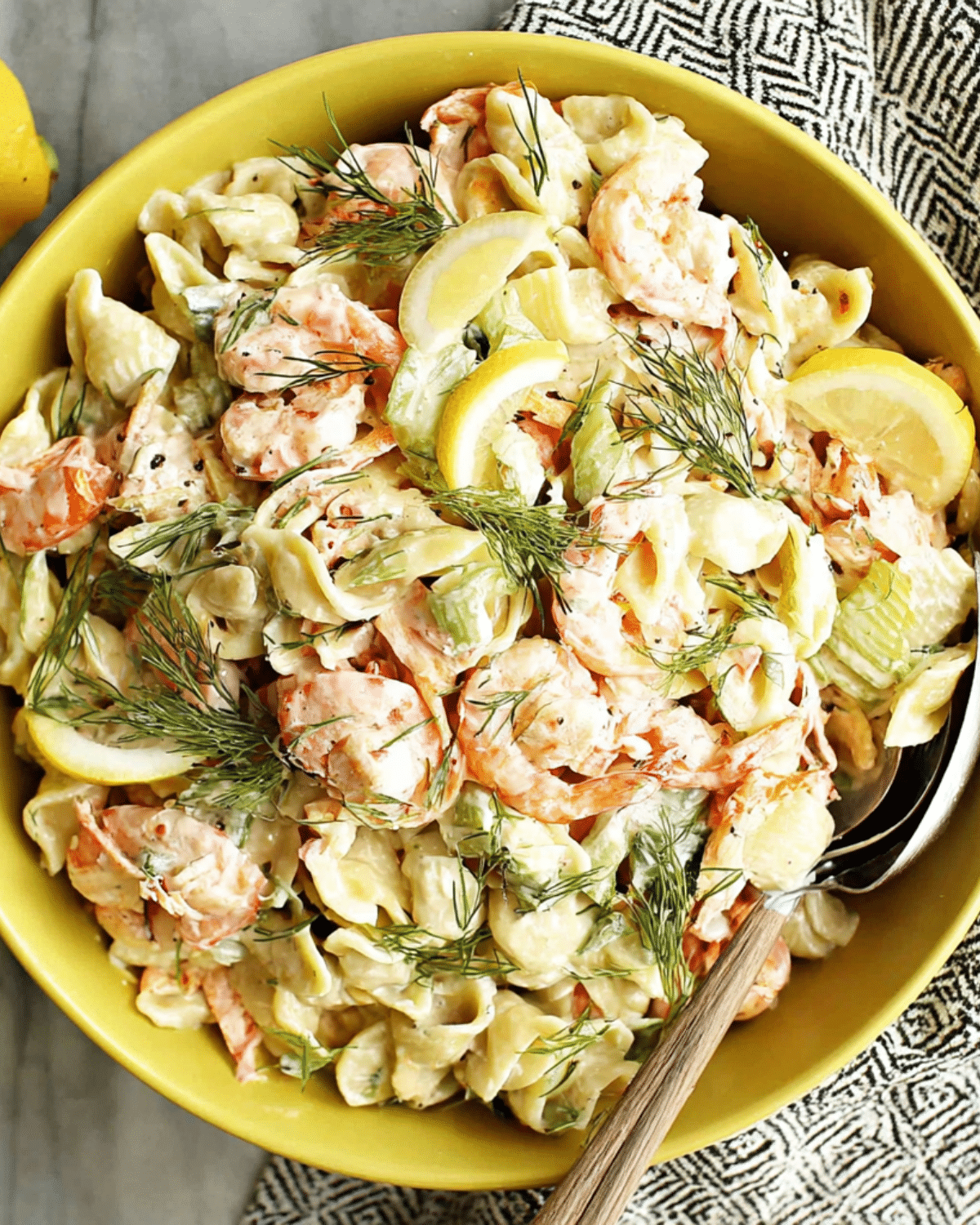 A bowl of creamy seafood pasta salad made with shell pasta, shrimp, celery, lemon slices, and fresh dill.