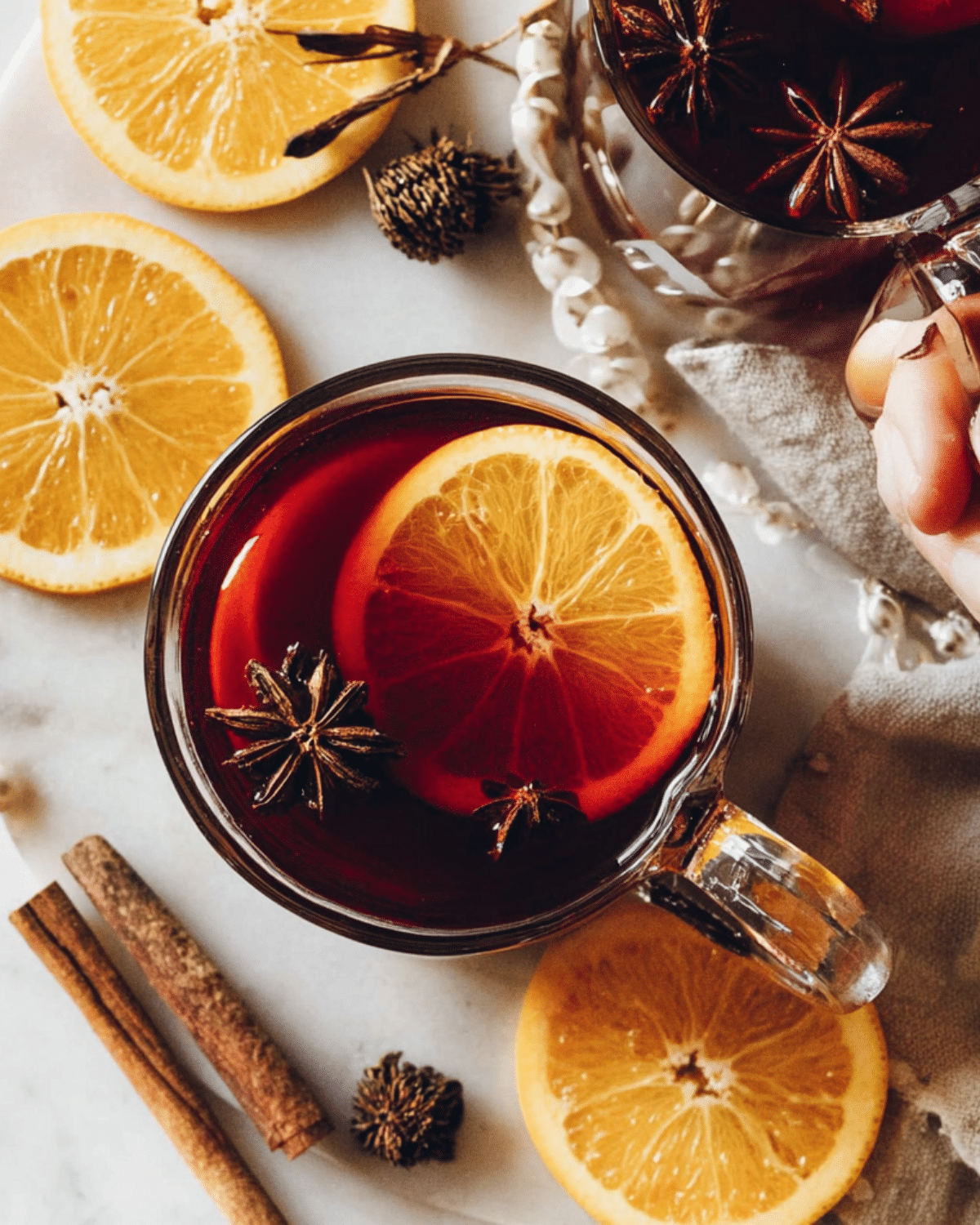 Glass mugs of tart cherry mulled drink garnished with orange slices, star anise, and cinnamon sticks on a marble surface.