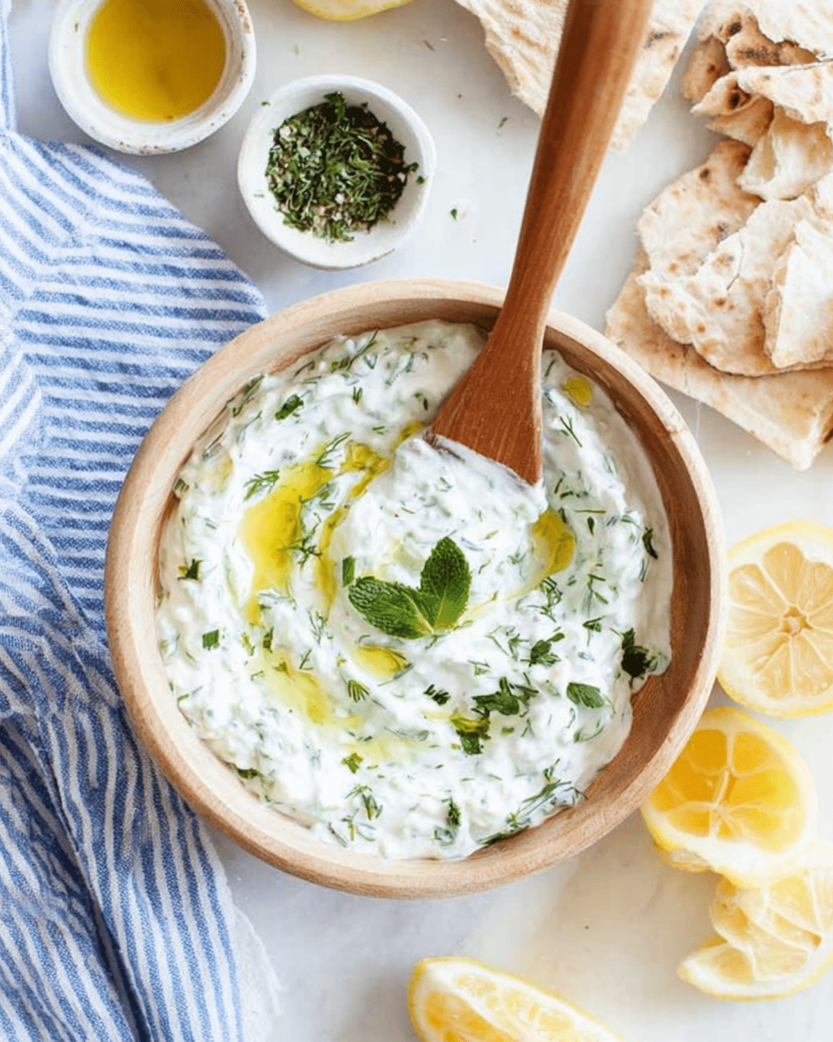 Healthy Tzatziki Sauce in a wooden bowl, topped with olive oil and fresh mint, surrounded by pita bread, herbs, and lemon slices.