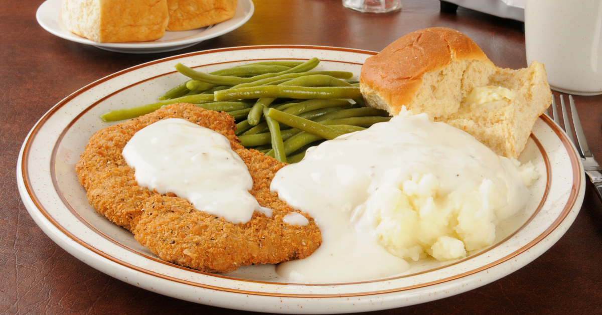 plate featuring chicken fried steak, accompanied by mashed potatoes, green beans, and a roll. This delicious and balanced meal is presented in a visually appealing manner.