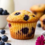 A golden, freshly baked blueberry muffin with plump blueberries peeking through, set against a light background.