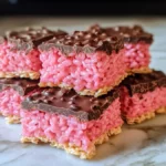 A stack of Chocolate Covered Cherry Rice Krispies Treats featuring a vibrant pink, cherry-flavored rice krispies layer topped with a thick coating of smooth chocolate. The treats are cut into neat squares, revealing the bright, chewy interior contrasted by the glossy chocolate topping. Placed on a marble surface, these colorful and delicious snacks offer a fun twist on the classic treat, perfect for dessert tables or special occasions.