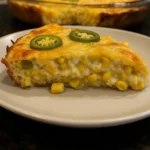 A thick slice of cheesy jalapeño corn bake on a plate, topped with fresh jalapeño slices and filled with whole golden corn kernels in a creamy cheese sauce.