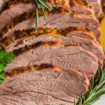 Sliced pork tenderloin roast garnished with fresh rosemary and parsley on a white platter.