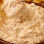 Creamy cheese dip with tomatoes and shredded cheese served in a brown bowl, surrounded by tortilla chips.