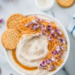 Overhead view of creamy dip topped with shredded cheddar cheese and diced red onion, surrounded by round crackers.