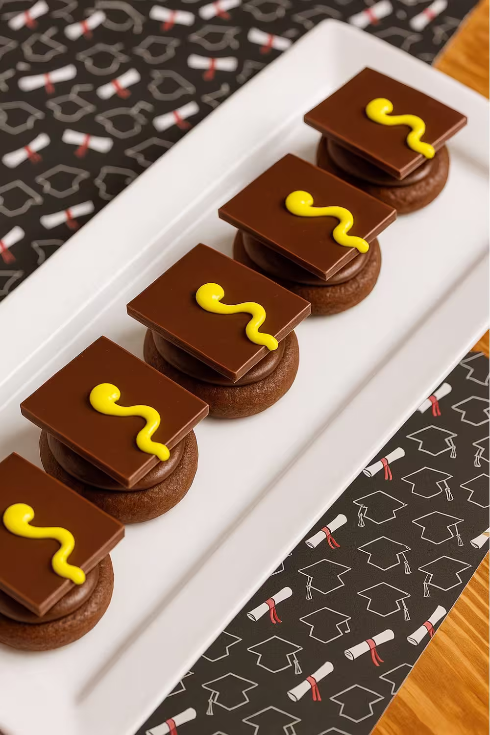 Chocolate cookies topped with a square chocolate piece resembling a graduation cap, complete with a yellow icing tassel, arranged neatly on a white platter.