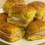 Stack of golden ham and cheese sliders topped with poppy seeds, served on a white plate.