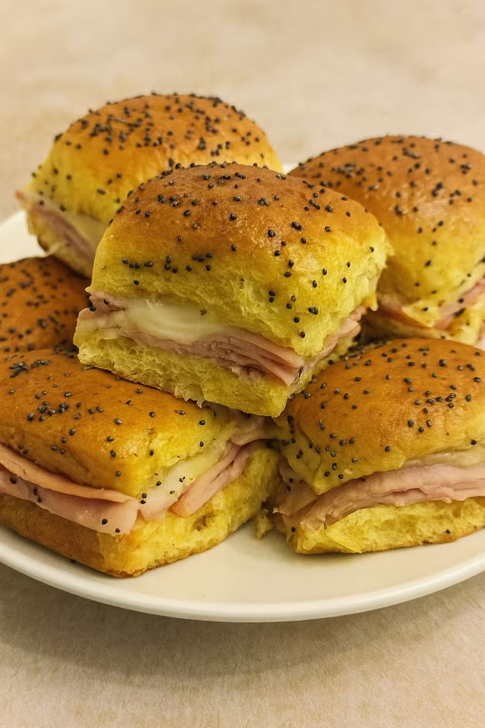 Stack of golden ham and cheese sliders topped with poppy seeds, served on a white plate.