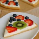Slice of gluten-free fruit pizza topped with fresh kiwi, strawberries, and blueberries on a creamy base.
