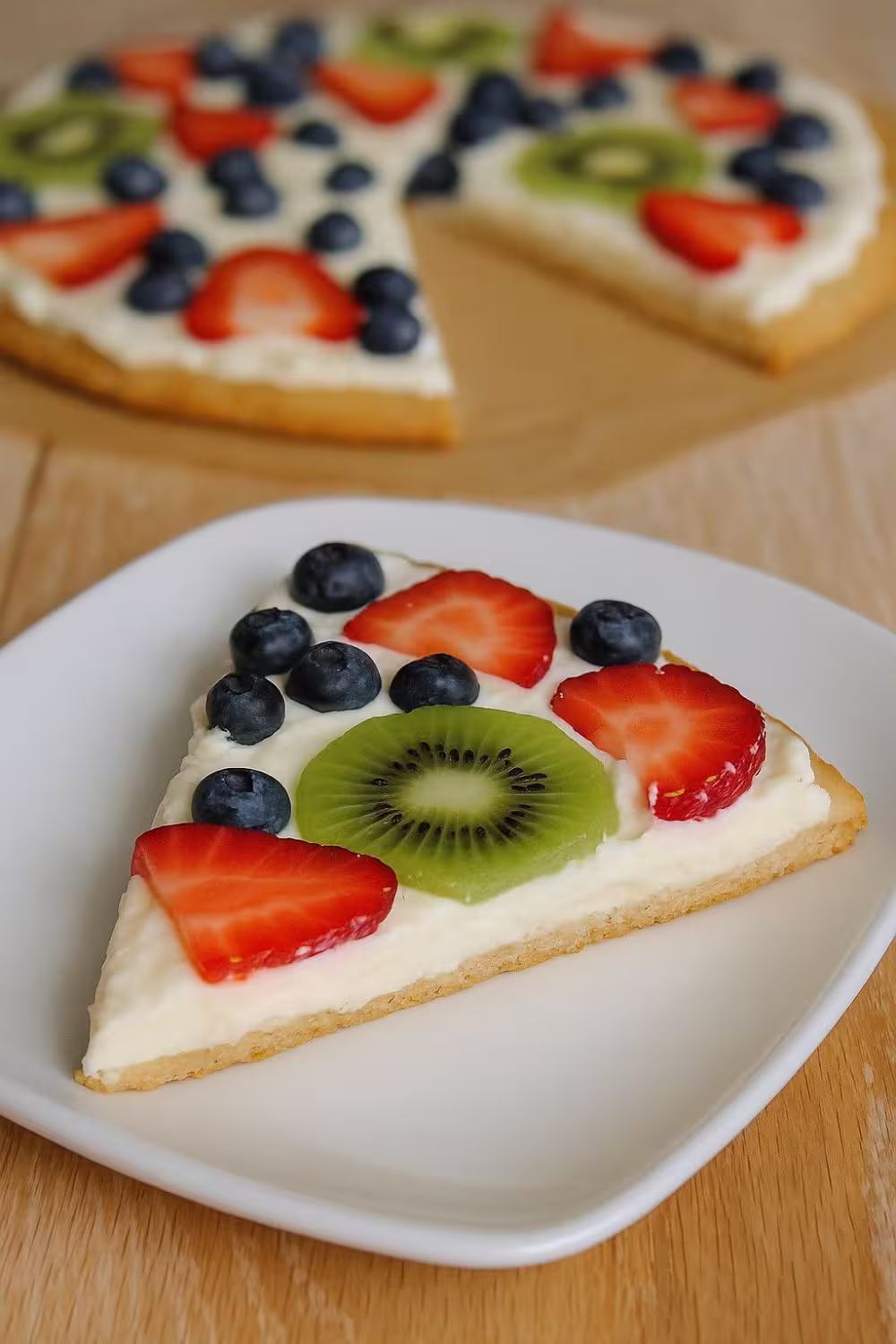 Slice of gluten-free fruit pizza topped with fresh kiwi, strawberries, and blueberries on a creamy base.