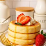 Stack of fluffy Japanese soufflé pancakes topped with whipped cream, fresh strawberry, and maple syrup.