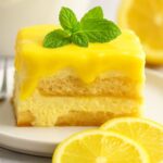 Slice of lemon tiramisu topped with lemon glaze and mint, served with fresh lemon slices.