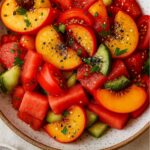 A colorful summer salad featuring fresh peach slices, juicy watermelon cubes, cucumber slices, fresh basil, and a sprinkle of black pepper, served in a rustic bowl.