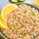 A bowl of creamy salmon salad garnished with fresh dill, red onions, and celery, served with lemon wedges for a refreshing flavor.