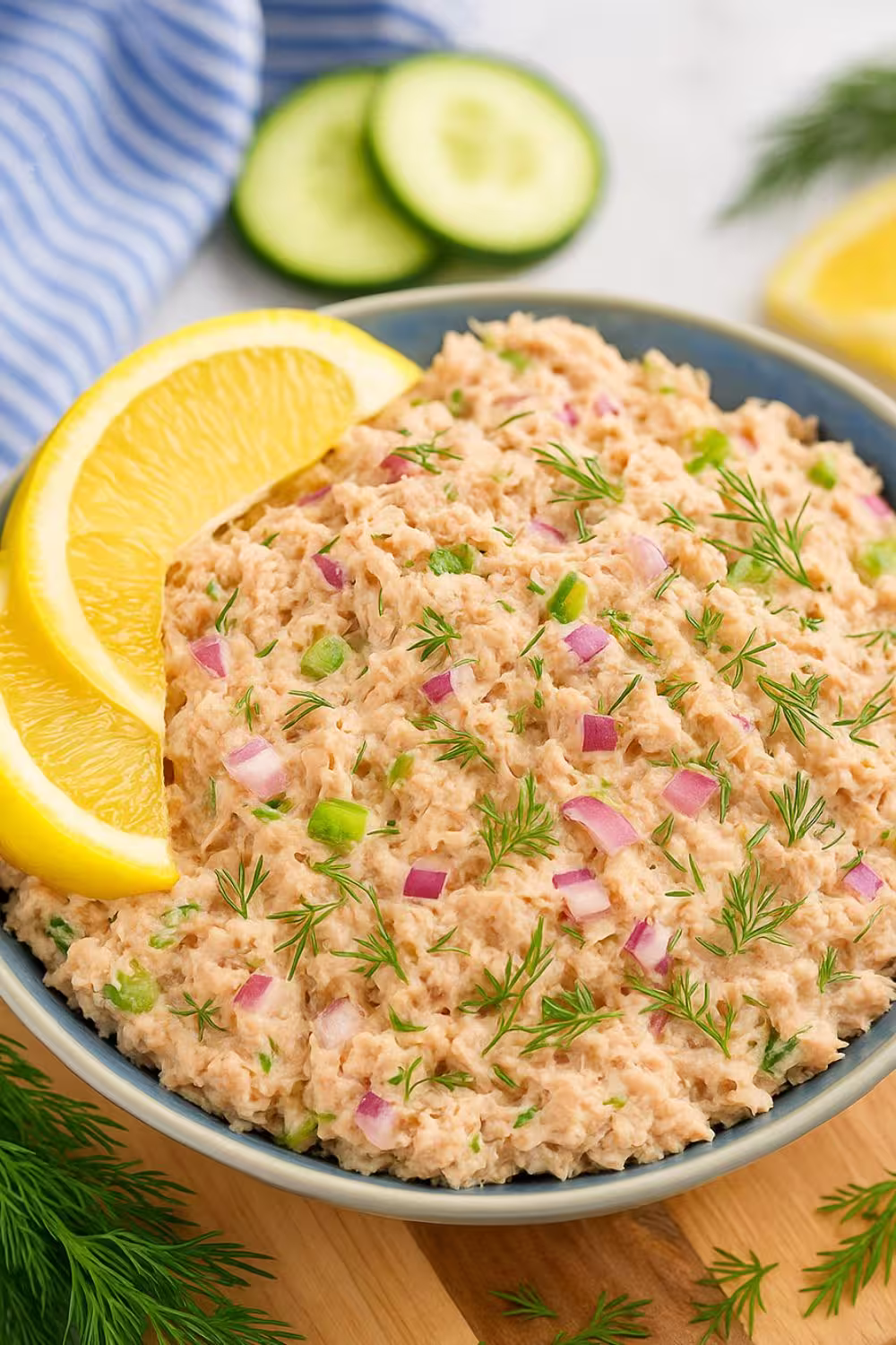 A bowl of creamy salmon salad garnished with fresh dill, red onions, and celery, served with lemon wedges for a refreshing flavor.