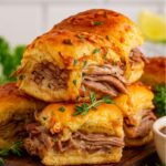 Stacked French Dip Sliders with tender roast beef, melted cheese, and savory au jus, garnished with fresh herbs on a rustic wooden board.