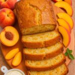 Sliced Southern Peach Bread on a wooden board, surrounded by fresh peach slices and a dollop of cream.
