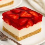 A square slice of Strawberry Icebox Cake featuring a crumbly graham cracker crust, creamy cheesecake layer, and a vibrant strawberry gelatin topping with fresh strawberry slices.