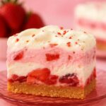 Close-up of a creamy No-Bake Strawberry Delight dessert slice on a pink glass plate, featuring layers of fresh strawberries, whipped cream, and a graham cracker crust.