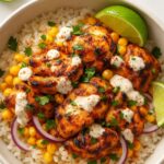 A flavorful Street Corn Chicken Rice Bowl topped with grilled chicken, charred corn, red onions, cilantro, lime wedges, and drizzled with creamy dressing, served with tortilla chips.