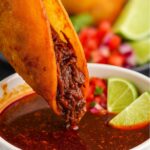 Crispy birria taco dipped into a rich, flavorful broth with lime wedges on the side.