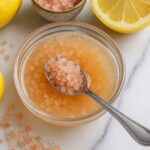 Japanese Pink Salt Recipe mixed with lemon juice, surrounded by lemons and a spoon scooping the salt.