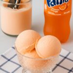 Scoops of creamy orange soda ice cream in a glass bowl with a bottle of Fanta and a tub of ice cream in the background.