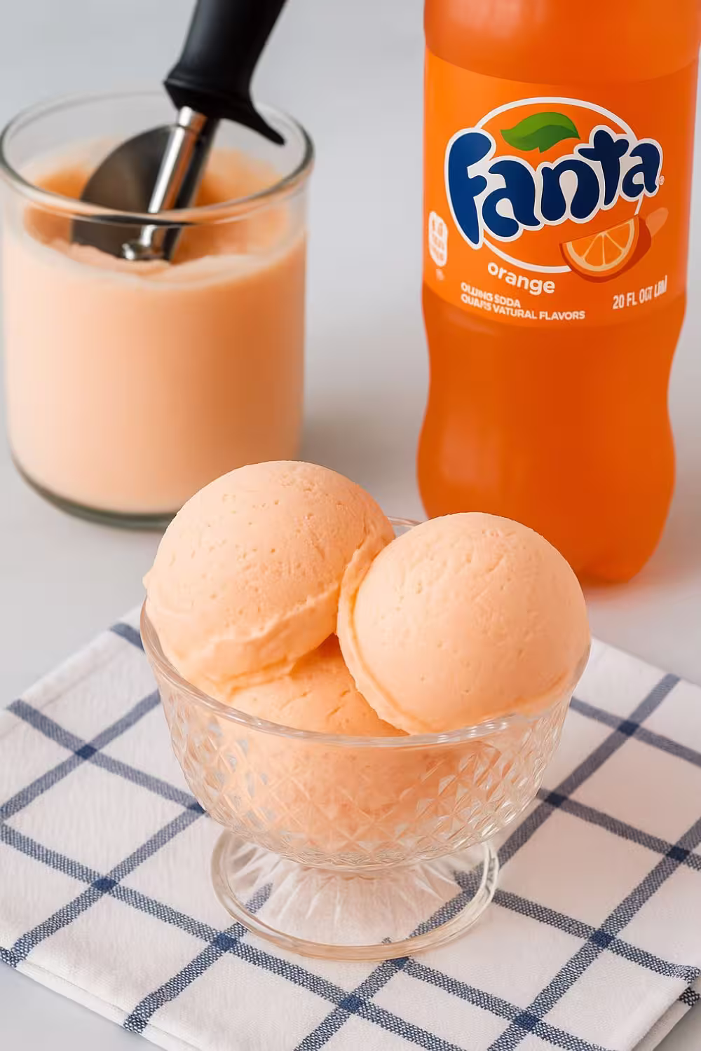 Scoops of creamy orange soda ice cream in a glass bowl with a bottle of Fanta and a tub of ice cream in the background.