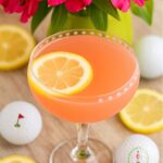 Refreshing Azalea drink garnished with a lemon slice in a decorative glass, surrounded by golf balls and bright pink flowers.