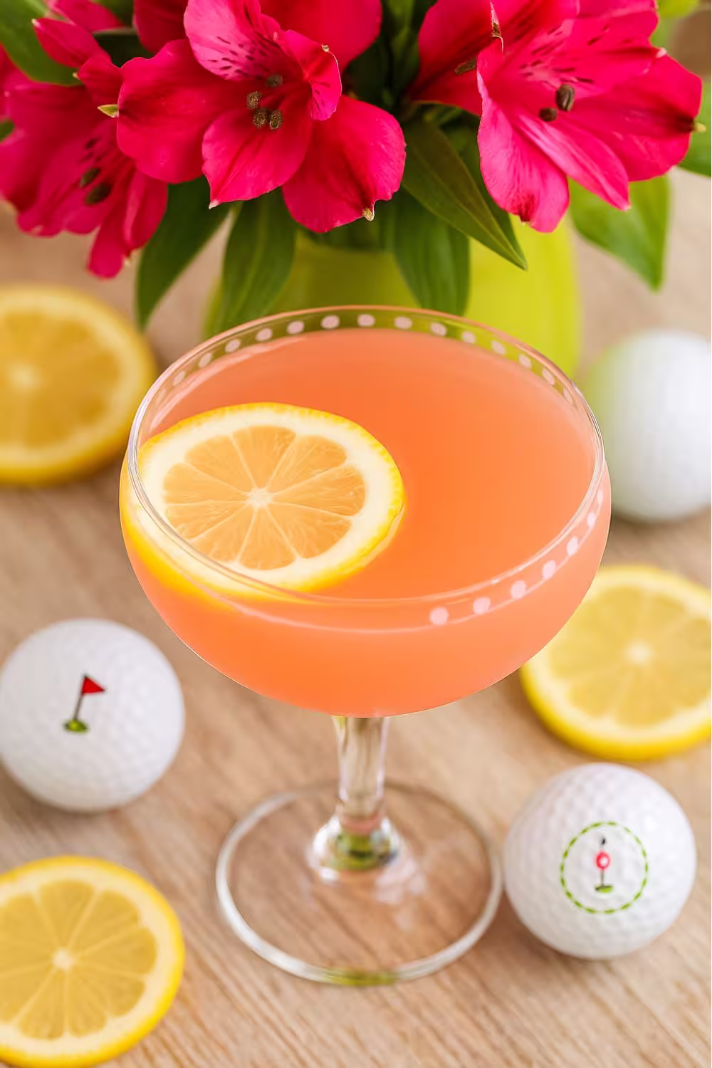 Refreshing Azalea drink garnished with a lemon slice in a decorative glass, surrounded by golf balls and bright pink flowers.
