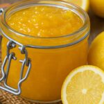 Glass jar filled with bright yellow Italian lemon jam, surrounded by fresh lemons and lemon halves on a rustic surface.