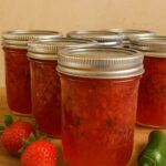 Canning jars filled with homemade strawberry jalapeño jam, surrounded by fresh strawberries and a green jalapeño pepper on a wooden surface.