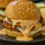 Close-up of a Bang Bang Chicken Slider with crispy chicken, melted cheese, and creamy sauce on a sesame seed bun.