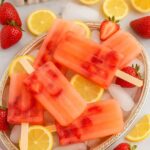 Refreshing Strawberry Lemonade Popsicles on a silver tray with ice, surrounded by fresh strawberries and lemon slices.