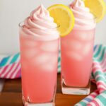 Two tall glasses of whipped pink lemonade topped with creamy pink whipped topping and lemon slices on the rim.