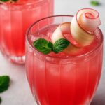 Refreshing pink rhubarb lemonade served in glass tumblers, garnished with mint leaves and a rhubarb curl.